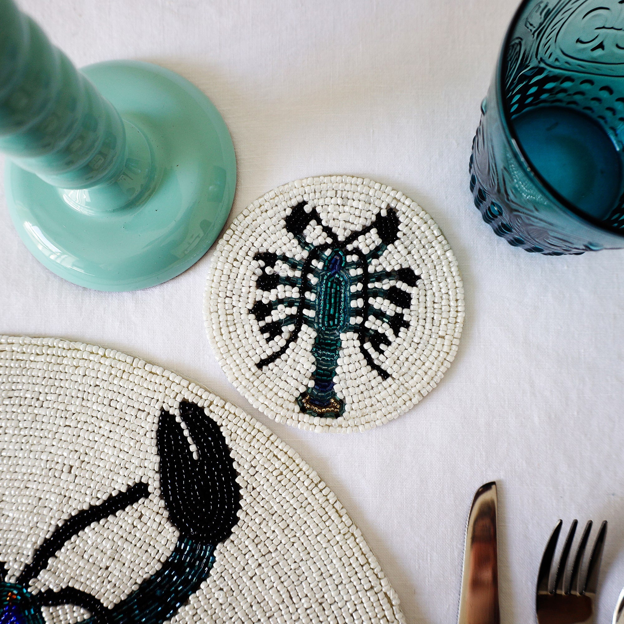 Handmade glass beaded coaster with a lobster design  next to a matching placemat on a table setting with glasses,cutlery and a candlestick