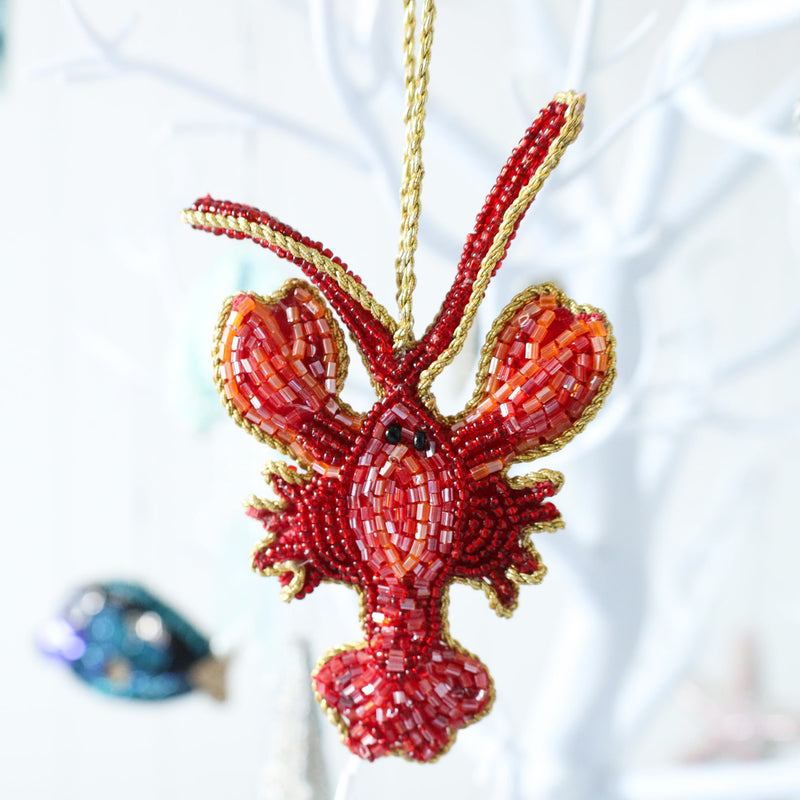 beaded read and orange lobster christmas decoration edged in gold braid hanging on a tree with the fish decoration in the background
