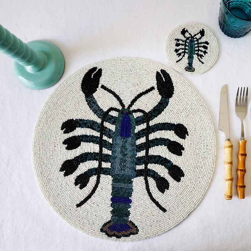 Handmade glass beaded coaster with a lobster design next to a matching placemat on a table setting with glasses,cutlery and a candlestick