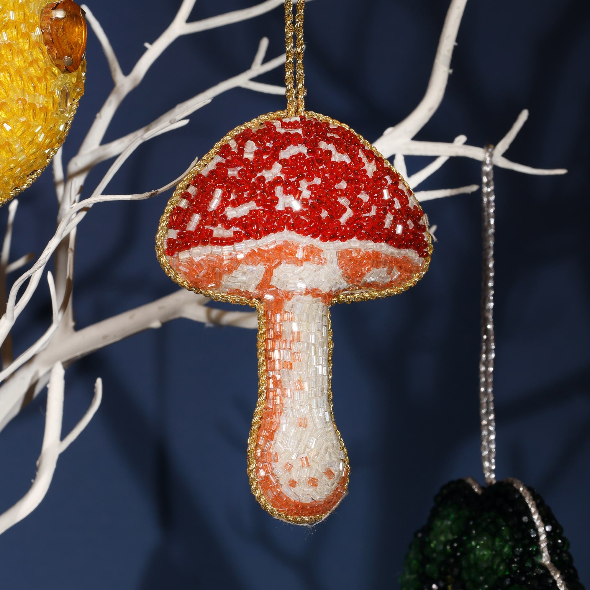 A detailed, beaded ornament shaped like a mushroom. The mushroom's cap is red with white spots, and the stem is a soft peach and white color, all outlined in gold beads. The intricate beading creates a textured and vibrant look, perfect for adding a whimsical touch to holiday decor. It hangs from a white decorative tree branch, adding an organic and festive charm.