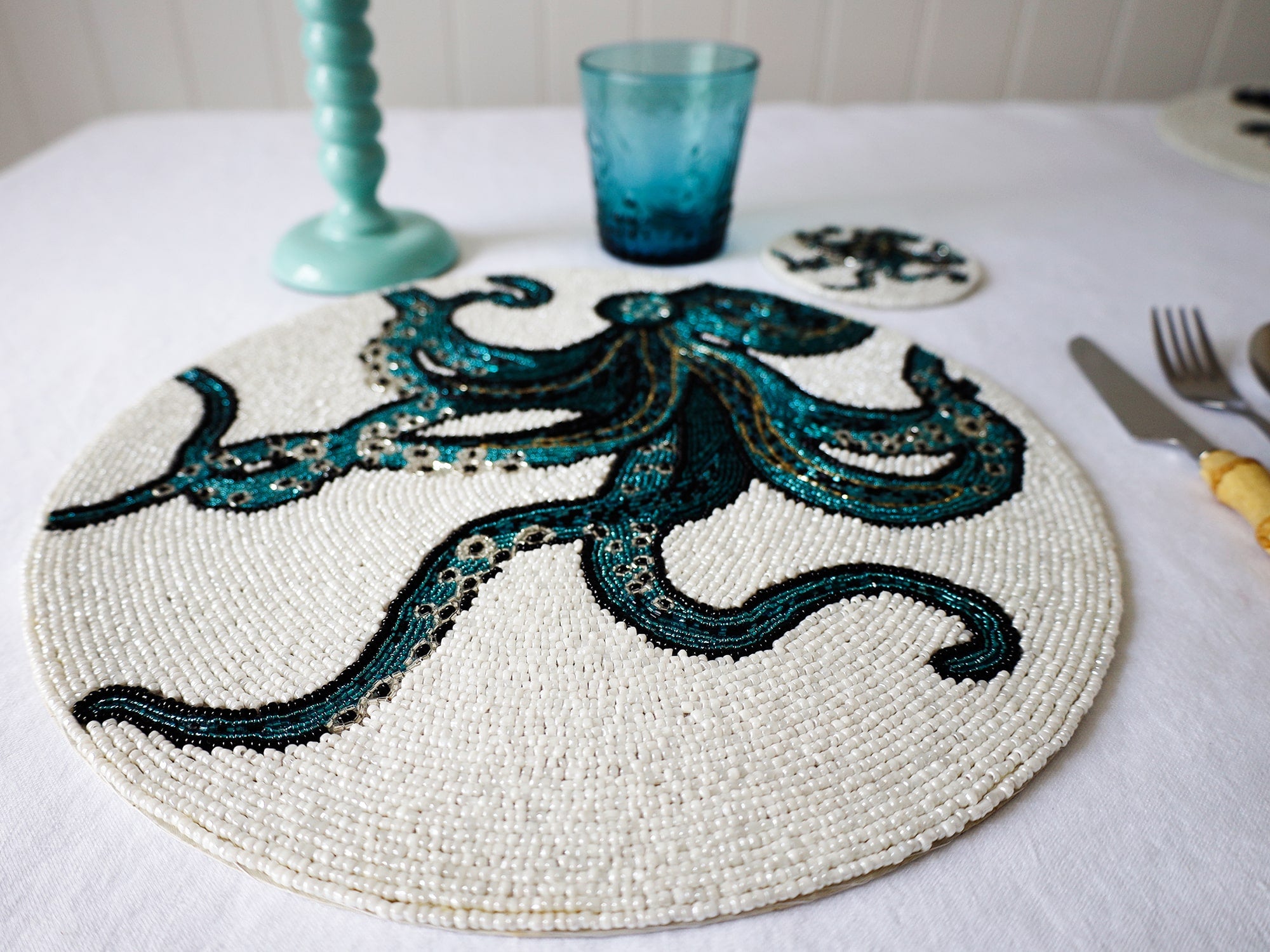 Handmade glass beaded placemat with a Octopus design in Navy,Black and gold on white glass beads in a place setting with cutlery,a glass and a candlestick