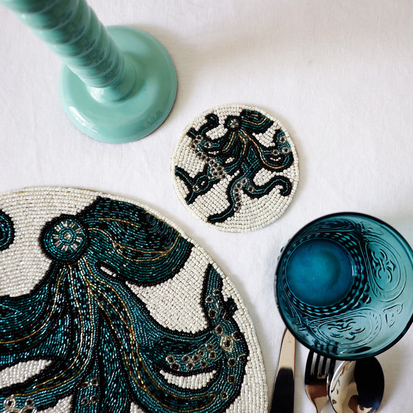 Handmade glass beaded coaster with an Octopus design next to a matching placemat on a table setting with glasses,cutlery and a candlestick