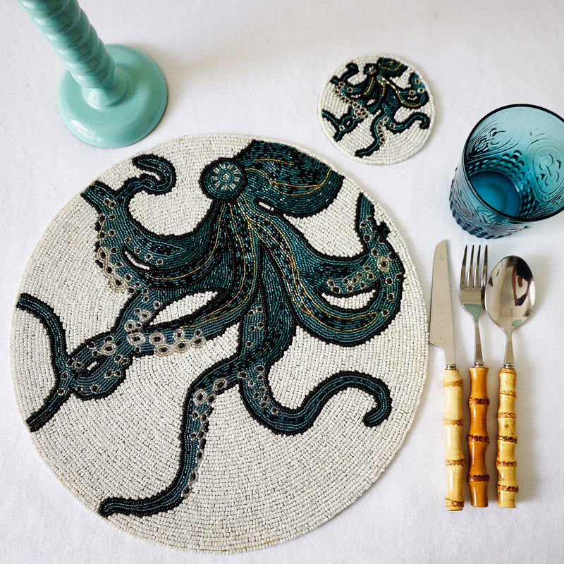 Handmade glass beaded coaster with an Octopus design next to a matching placemat on a table setting with glasses,cutlery and a candlestick
