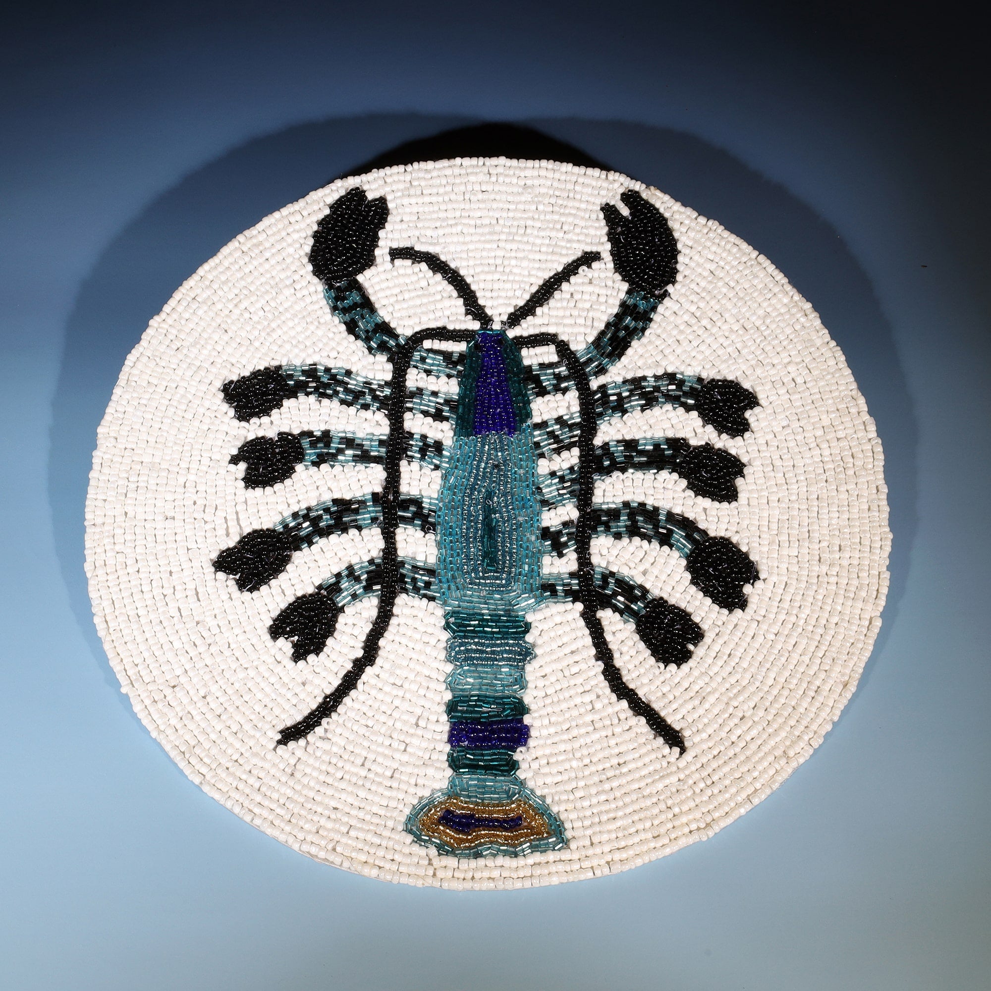 The image shows a round beaded placemat featuring a detailed design of a lobster. The lobster is depicted in shades of teal, black, and blue, with intricate beadwork that highlights its segmented body, claws, and antennae. The design is bold and symmetrical, with the lobster's vibrant colors standing out against the neutral white beaded background. The craftsmanship of the beadwork adds texture and dimension to the piece, making it a striking and artistic representation of marine life