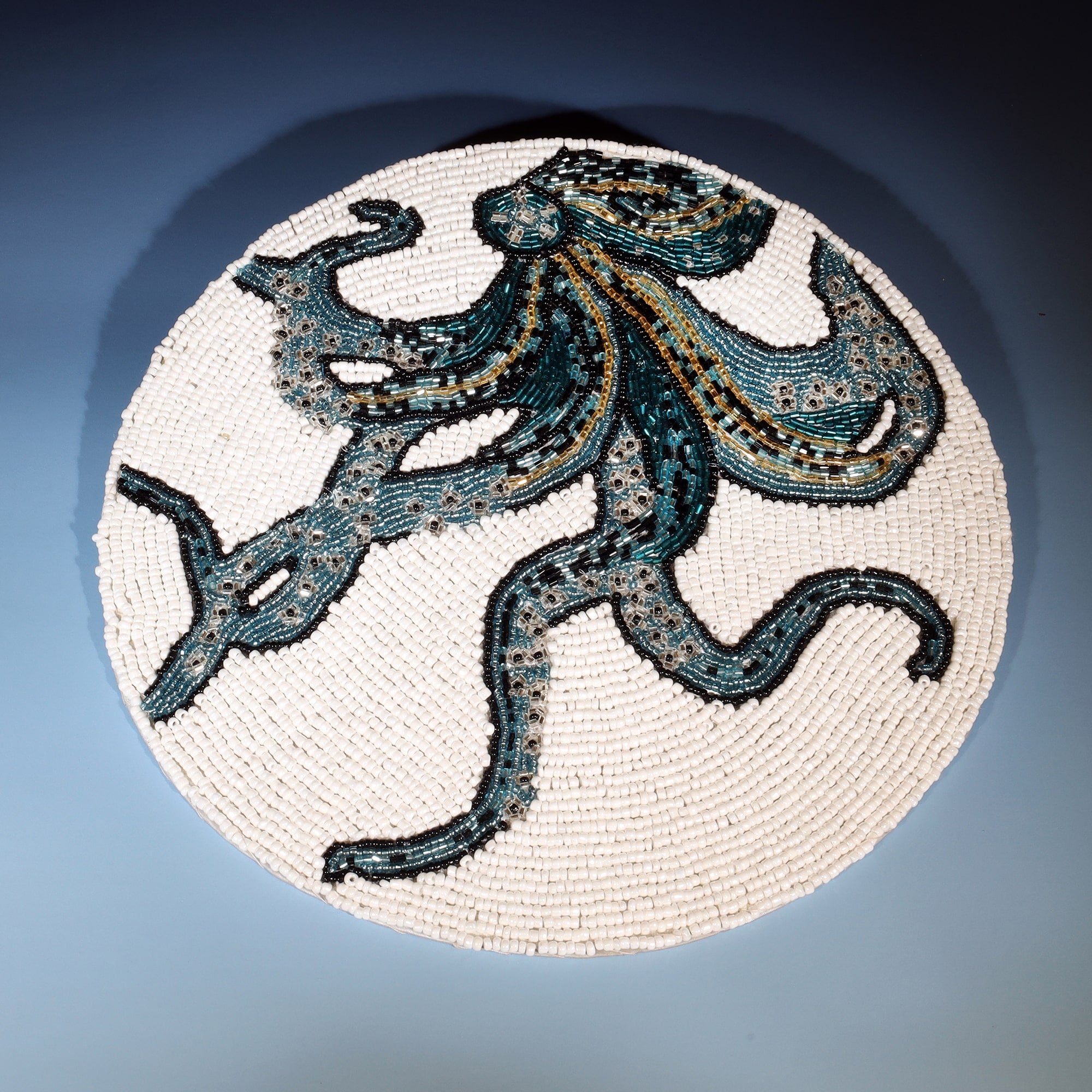 The image shows a round beaded placemat featuring a detailed illustration of an octopus. The octopus is depicted in shades of teal, navy blue, and gold, with intricate beadwork creating a textured and vibrant design. The background of the placemat is made up of small white beads, which contrast beautifully with the colorful octopus, highlighting its swirling tentacles and flowing form. The craftsmanship of the beadwork adds depth and dimension to the overall piece, making it a striking and artistic represen