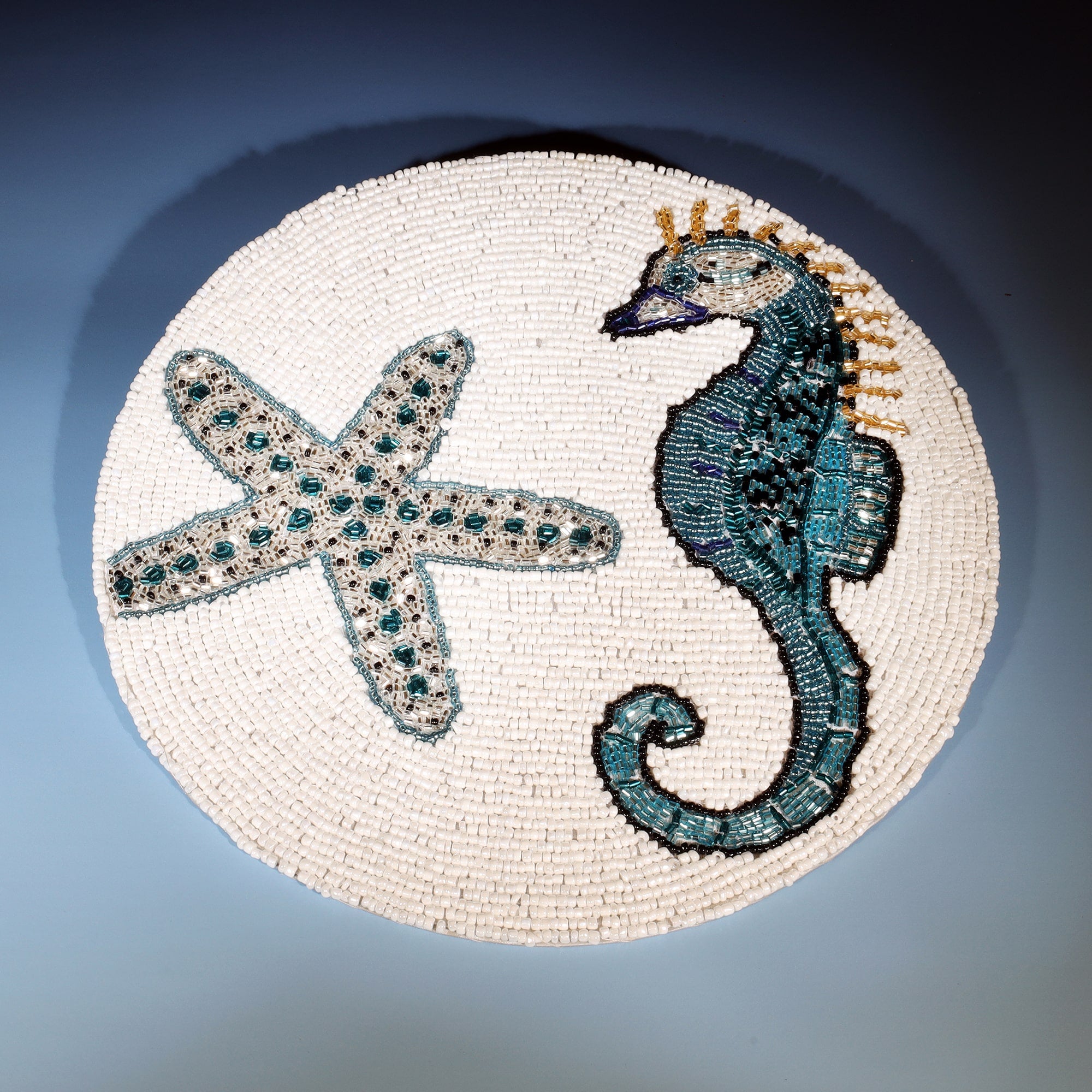 The image shows a round beaded placemat featuring a detailed design of a seahorse and a starfish. The seahorse is depicted in shades of teal, blue, and black, with intricate beadwork that highlights its curved body and crown-like fin. The starfish, positioned beside the seahorse, is adorned with teal and silver beads, creating a textured and sparkling effect. The background of the placemat is made up of small white beads, allowing the marine creatures to stand out with vibrant contrast. The craftsmanship an