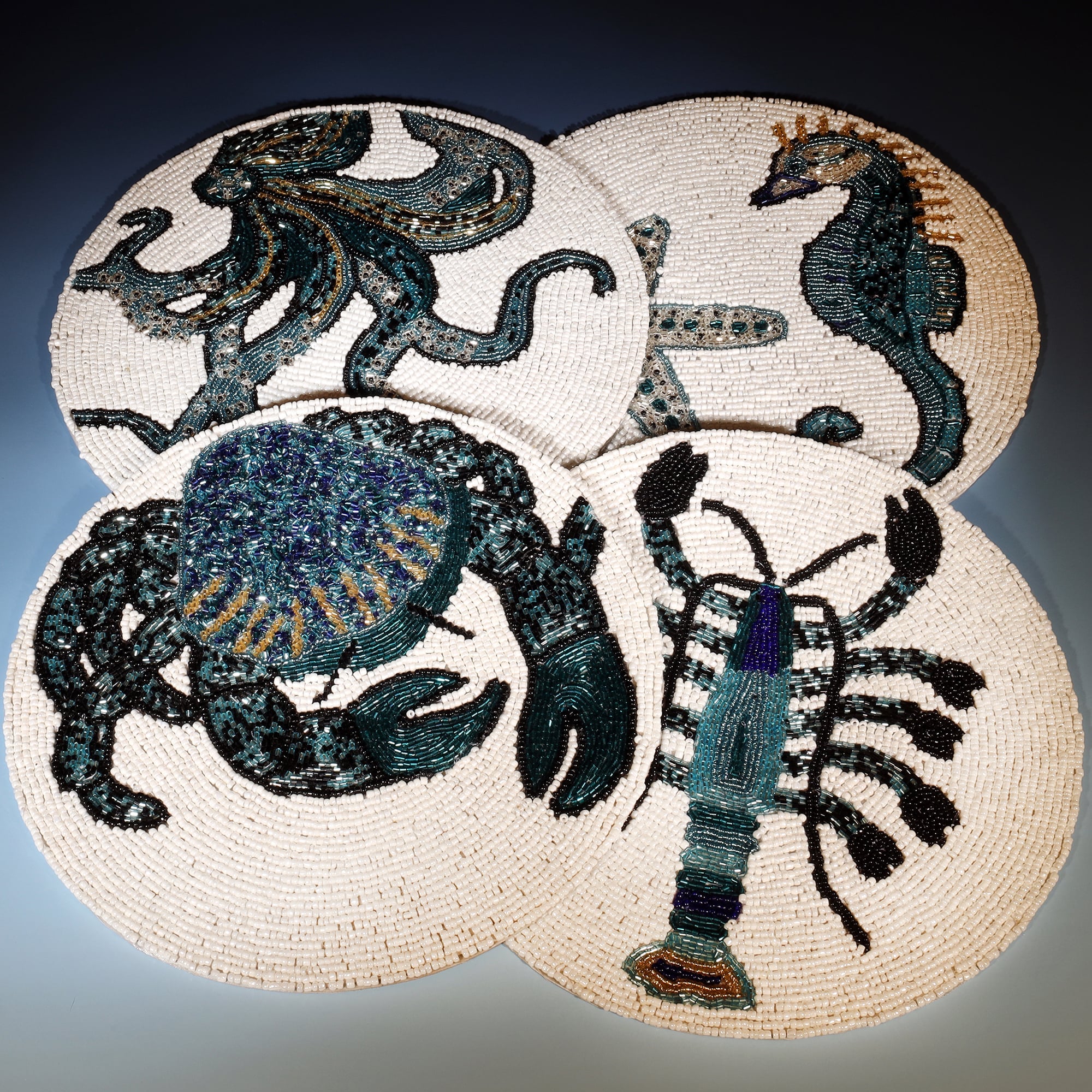 The image shows a set of intricately beaded placemats featuring marine life designs. Each circular placemat is decorated with a detailed depiction of a different sea creature, including an octopus, seahorse, crab, and lobster. The designs are rendered in shades of navy blue, teal, and black, with the beadwork creating a textured and vibrant appearance against a neutral background. The placement of the placemats creates a visually appealing arrangement that highlights the artistry and craftsmanship of the be