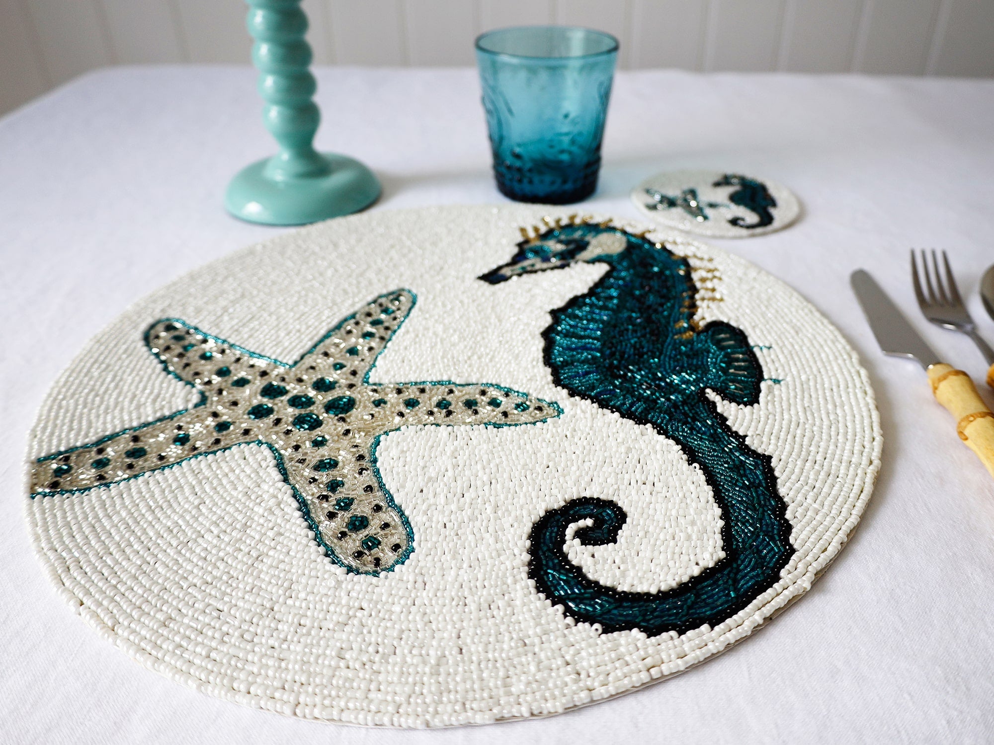Handmade glass beaded placemat with a Seahorse and Starfish design in Navy,Black and gold on white glass beads in a place setting with cutlery,a glass and a candlestick