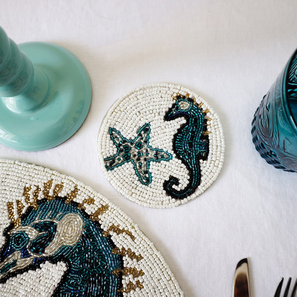 Handmade glass beaded coaster with a seahorse and starfish design next to a matching placemat on a table setting with glasses,cutlery and a candlestick