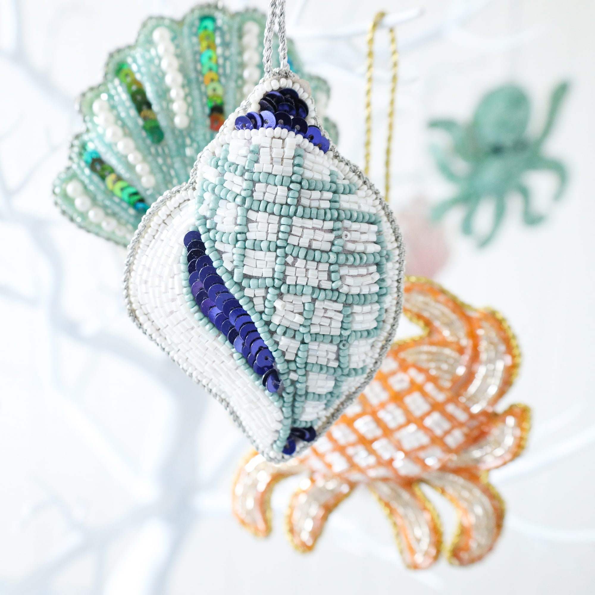 beaded blue and white christmas decoration in the shape of a conch shell hanging on a tree with an aqua scallop shell and a gold crab  in the background