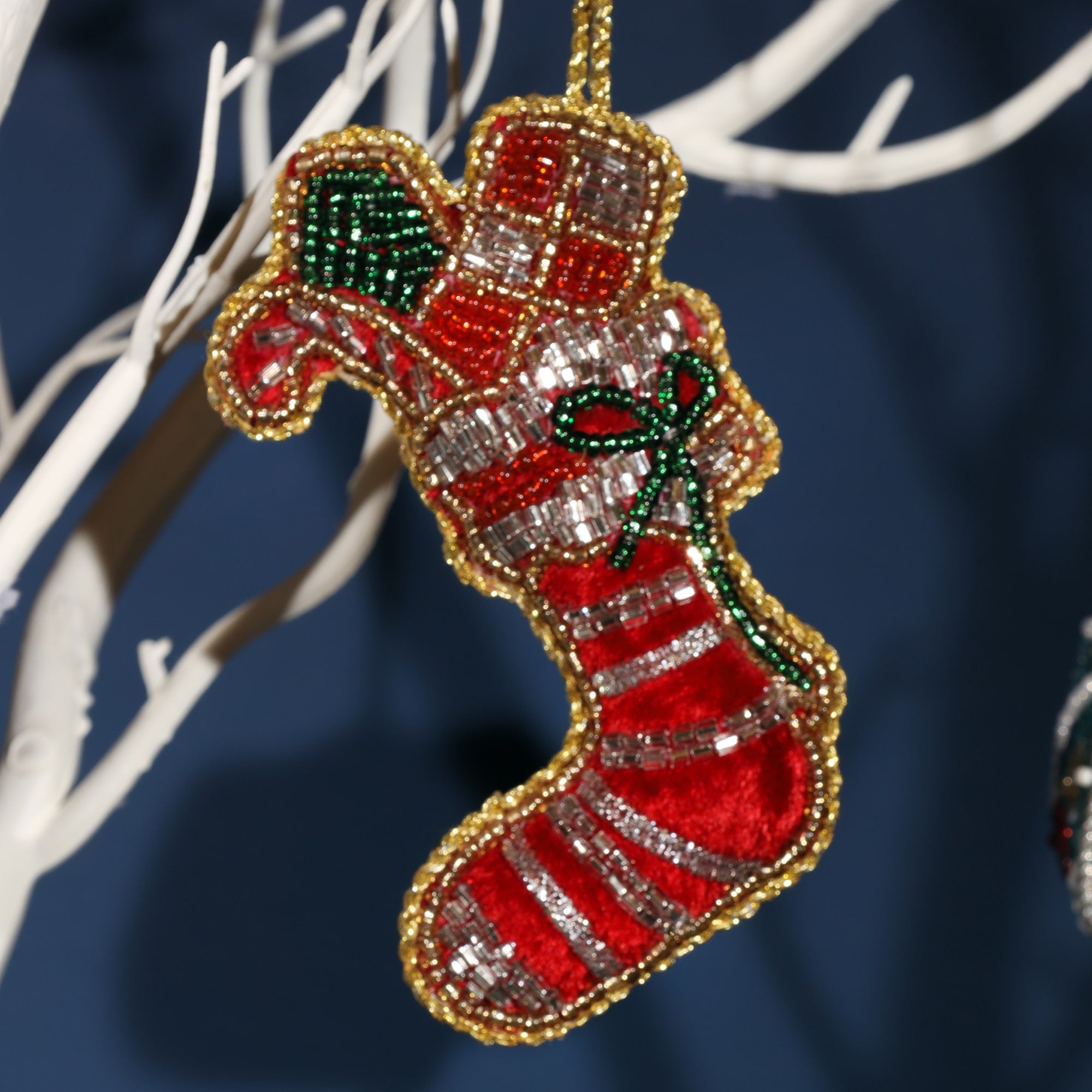 A festive beaded Christmas stocking ornament, decorated with red and white stripes, silver beads, and a small green bow. The ornament is outlined with gold beads, hanging from a white branch against a dark background.