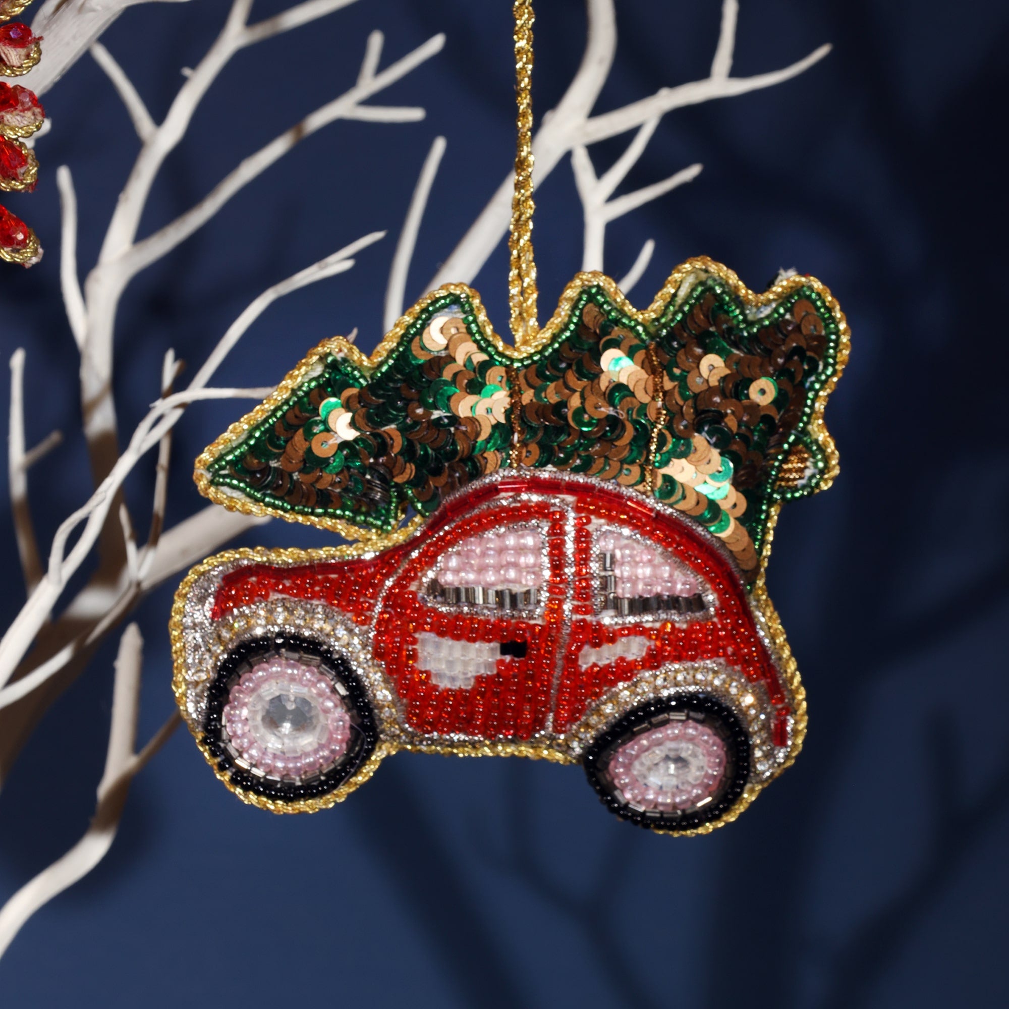 A detailed, beaded ornament of a red car carrying a Christmas tree on its roof. The car is intricately designed with red, silver, and black beads, and the windows are adorned with shimmering pink beads. The tree on top is decorated with green and brown sequins, giving it a festive sparkle. The ornament is bordered with gold beads, adding an elegant touch to the playful holiday theme.