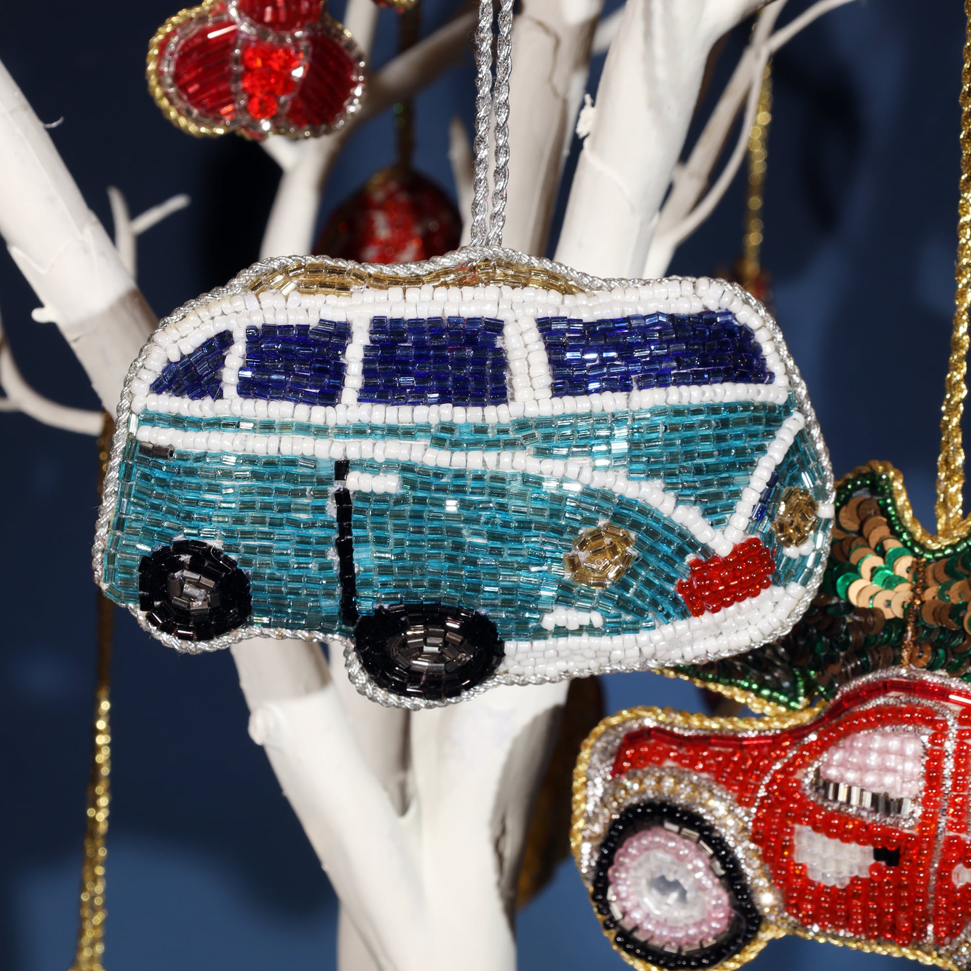 A close-up of a beaded ornament shaped like a blue and white VW camper van, hanging from a white branch. The ornament is intricately detailed with tiny blue, white, and silver beads, with black wheels and a red bumper. In the background, other colorful beaded decorations, including a red car, add to the festive display.