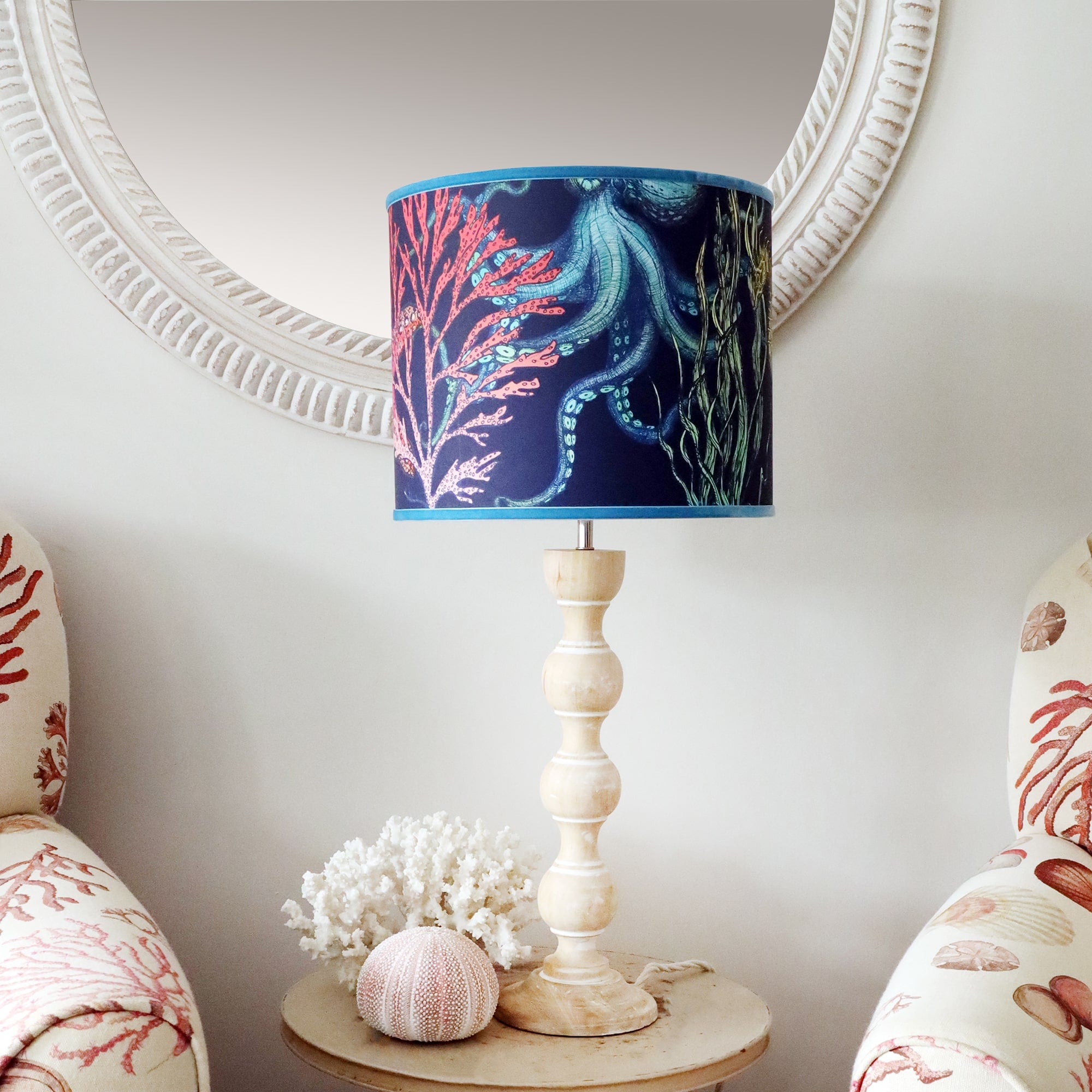 Whitewashed wooden bobbin lamp base on side table with rainbow reef navy lampshade, sitting between two coastal chairs and a whitewashed wooden mirror hanging on the wall behind.