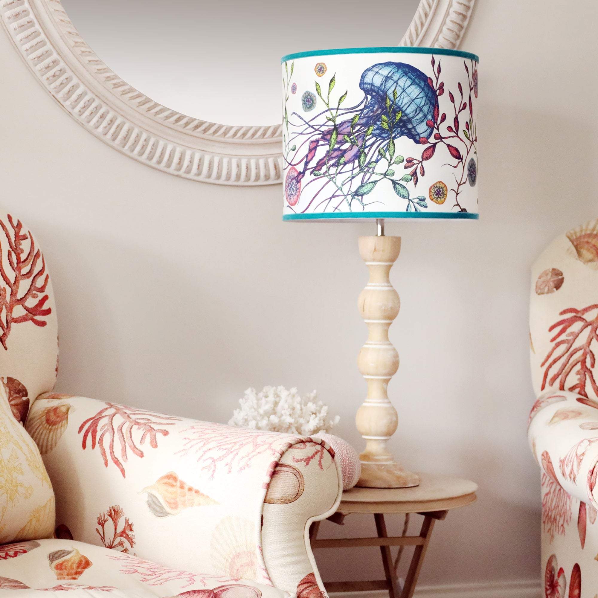 Whitewashed wooden bobbin lamp base on a metal side table with jellyfish reef lampshade, sitting between two coastal arm chairs and a whitewashed rope effect mirror hanging on the wall behind.