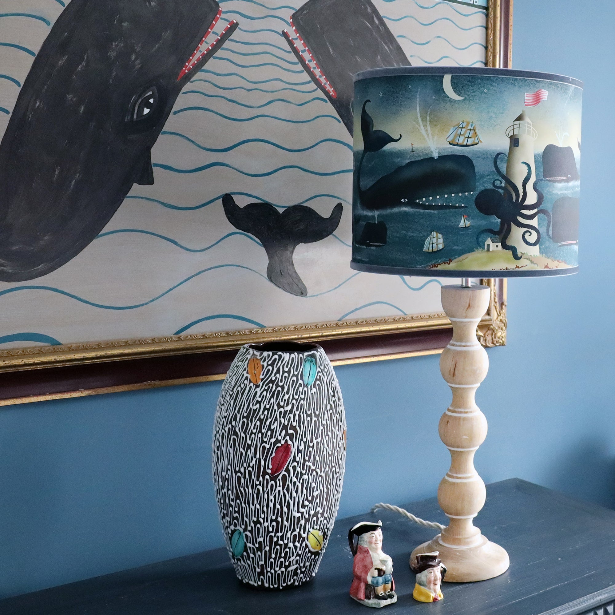 Whiewashed wooden bobbin lamp base on chest of drawers with whale & kraken lampshade and a large naive whale painting hanging on the wall behind.