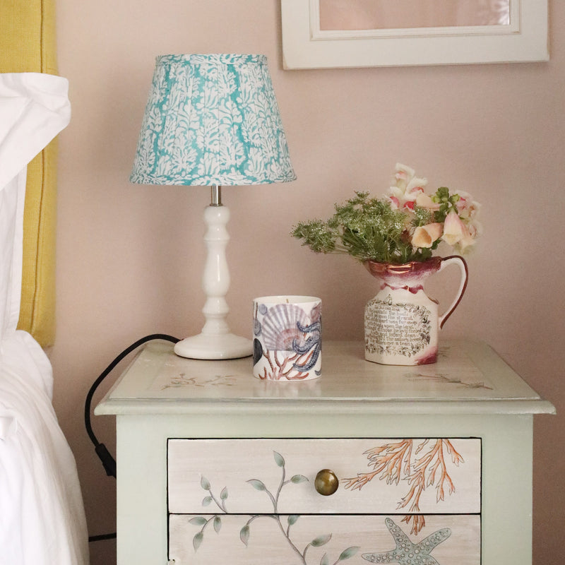 Small pleated Summer Skies Coraline pleated lampshade in hand blocked print on a white lampbase on a bedside table.Also on the table is a beachcomber candle and a jug with flowers
