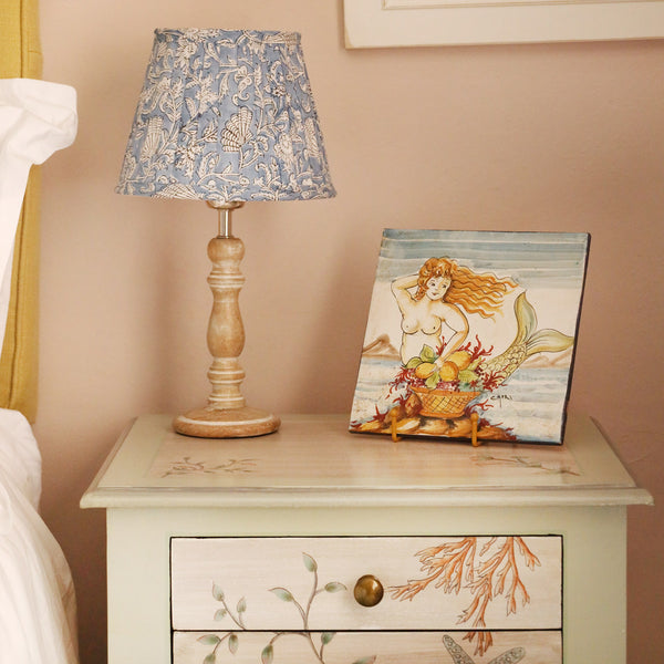 Small pleated Atlantic Blue Seashell lampshade in hand blocked print in blue and white on a wooden lampbase placed on our hand painted bedside table.On the table is a painting of a mermaid