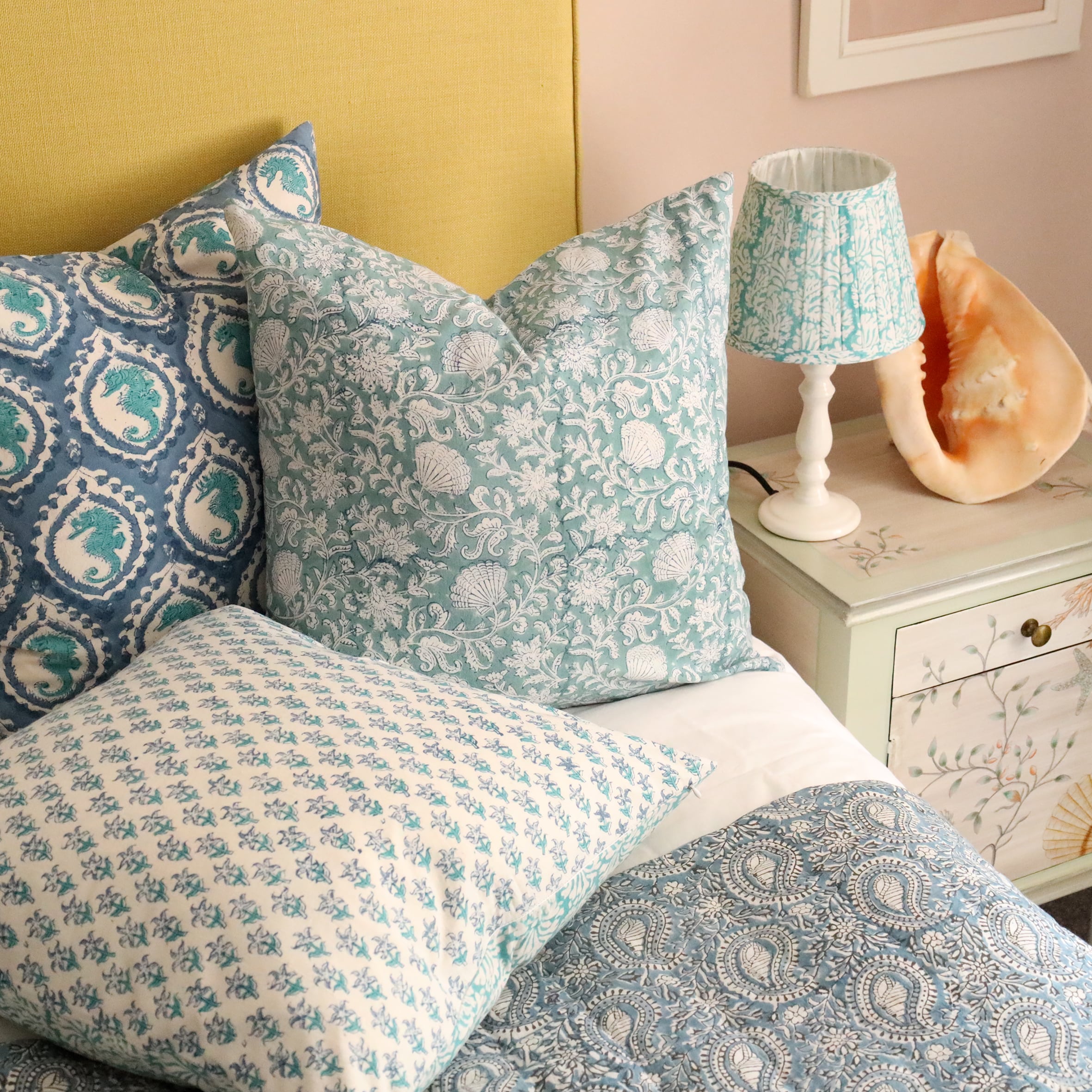 Close up of Sea Breeze Seashell Flower cushion which is Hand block printed fabric in a turquoise blue .The cushion is placed on a bed with 2 other block print cushions behind in on a crisp white bed,in front of a mustard yellow headboard.Next to the bed is a white lamp base with a pleated block print fabric lampshade on a hand painted bedside table