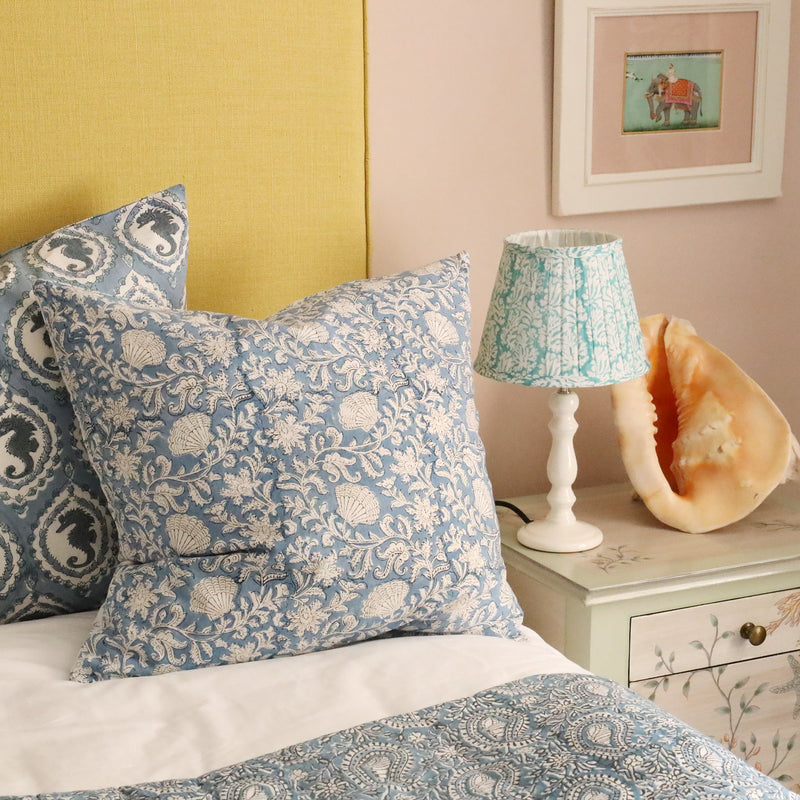 Small pleated Summer Skies Coraline pleated lampshade in hand blocked print on a white lampbase on a bedside table,also on the table is a large shell.In the foreground you can see our new bed linen displayed on a bed with several cushions