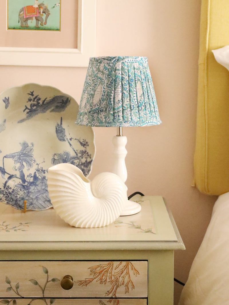 Small pleated Seafoam Paisley Shell lampshade on a white lampbase.The lampbase is on a hand painted bedside cabinet,also on the cabinet ia a large white shell and a blue and white decorated plate.On the wall behind you can see a painting of a man on an elephant