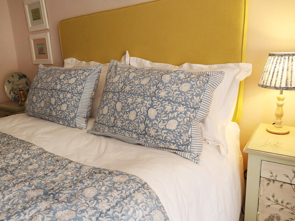 Two of the Seashell Flower pillowcases which in a Hand block printed fabric in a soft blue and white.They are filled with inners and placed on top of two white pillows on a  white bed with one of our matching quilts in the foreground.The headboard of the bed in a mustard yellow, on either side are bedside cabinets decorated with lamp bases and other decorations