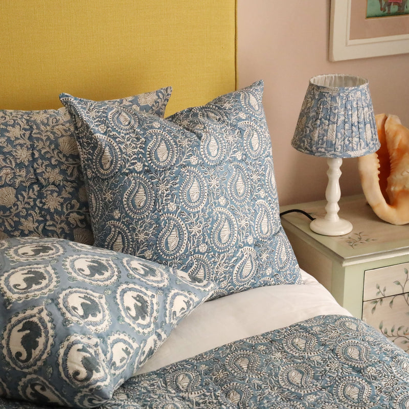 Close up of Azure Paisley Shell cushion .The cushion is placed on a bed with 2 other block print cushions behind in on a crisp white bed,in front of a mustard yellow headboard.Next to the bed is a cream lampbase with a pleated block print fabric lampshade on a hand painted bedside table