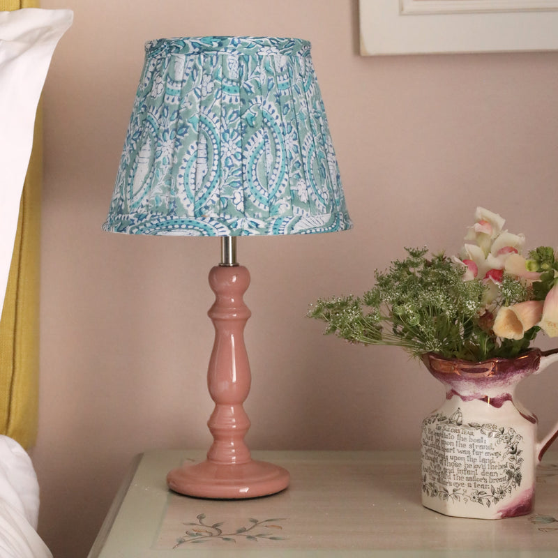 Blush pink lacquered finish wood lamp base with a pleated 20cm lampshade on a hand painted bedside cabinet.on the cabinet is a small decorated jug with flowers in