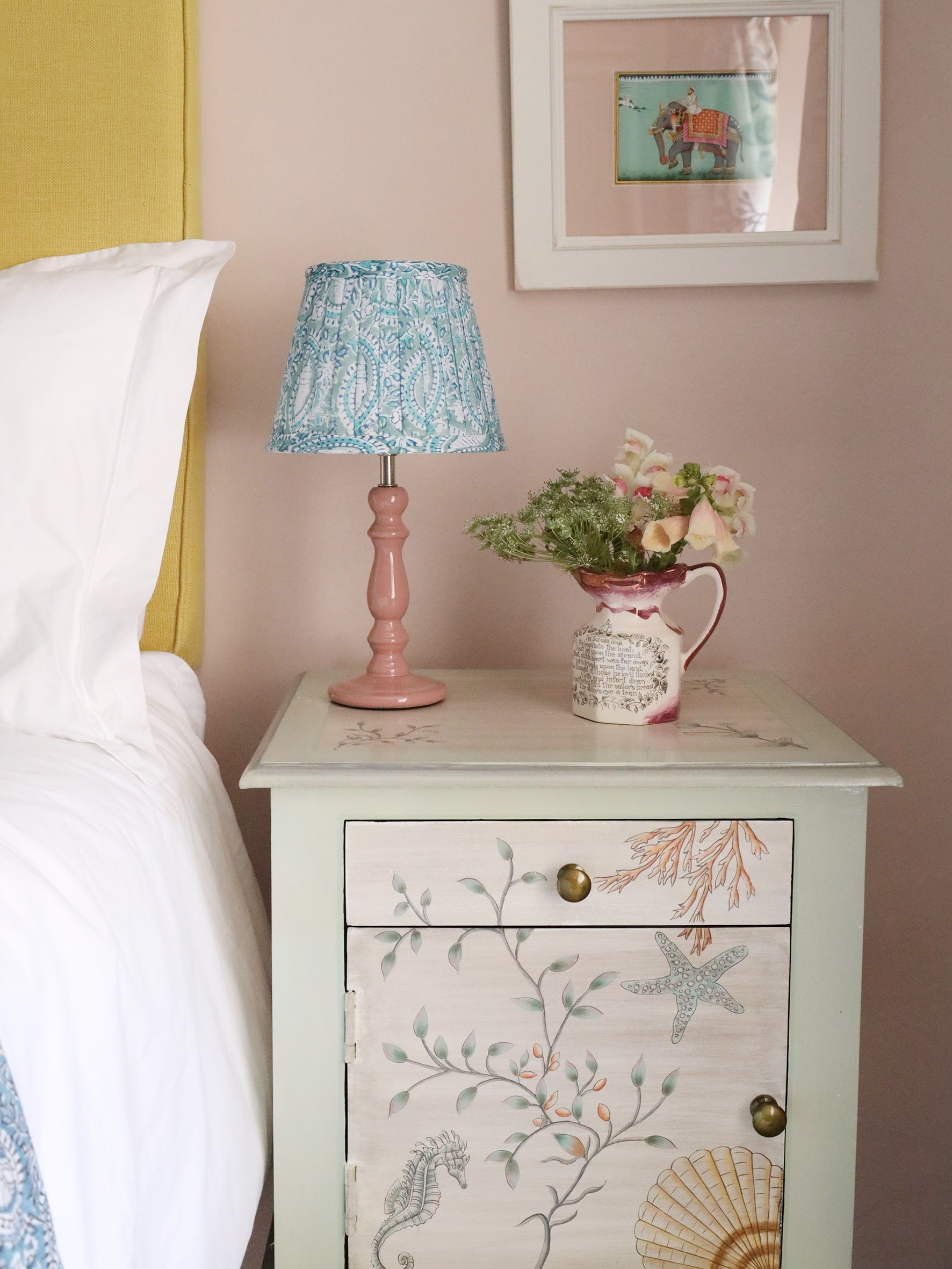 Small pleated Seafoam Paisley Shell lampshade in hand blocked print on a pink lampbase.The lampbase is on a bedside cabinet with a small jug with flowers.It is next to a bed,on the wall is a painting of a man on an elephant