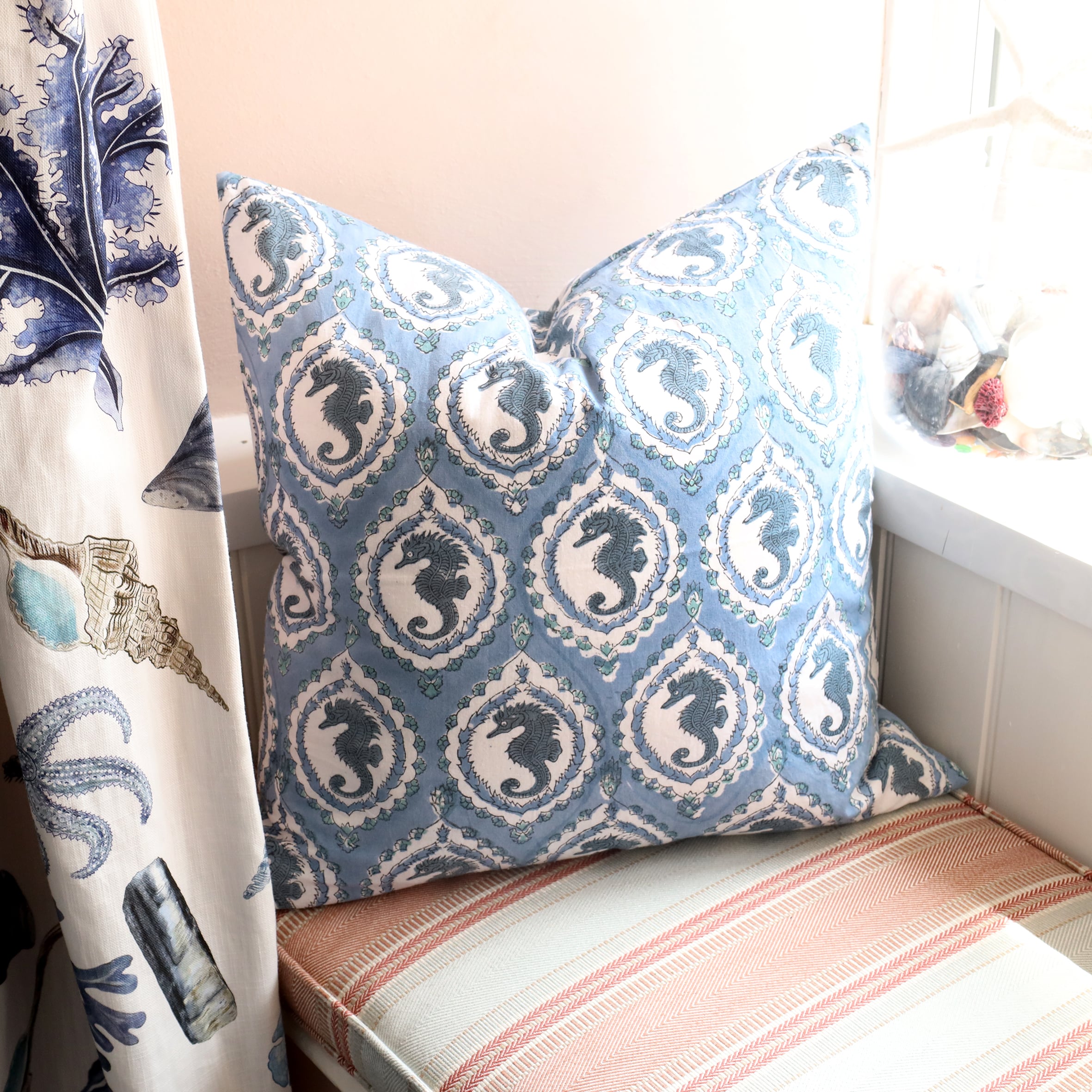 Ocean Blue Seahorse cushion which is Hand block printed fabric in a  blue and navy seahorse in a cameo design on a window seat.On the window ledge is a jar of shells on the window ledge