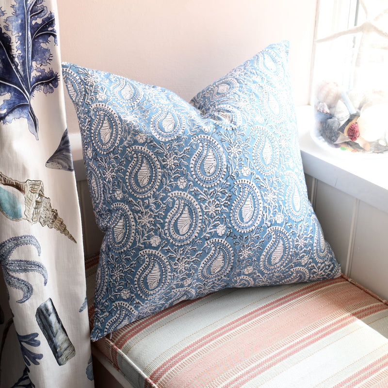 Azure Paisley Shell cushion which is Hand block printed fabric in a soft blue and white on a window seat.You can see the Rockpool curtain in the foreground and a glass jar with shells in.