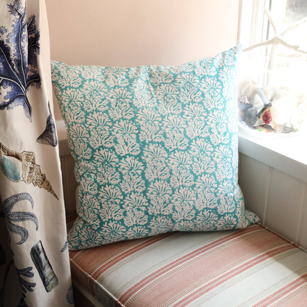 Coastal Blue Coraline cushion which is Hand block printed fabric in a soft turquoise blue with continuous white coral print.Cushion is on a window seat setting with a curtain made in our Rockpool fabric and a jar of shells on the window ledge.