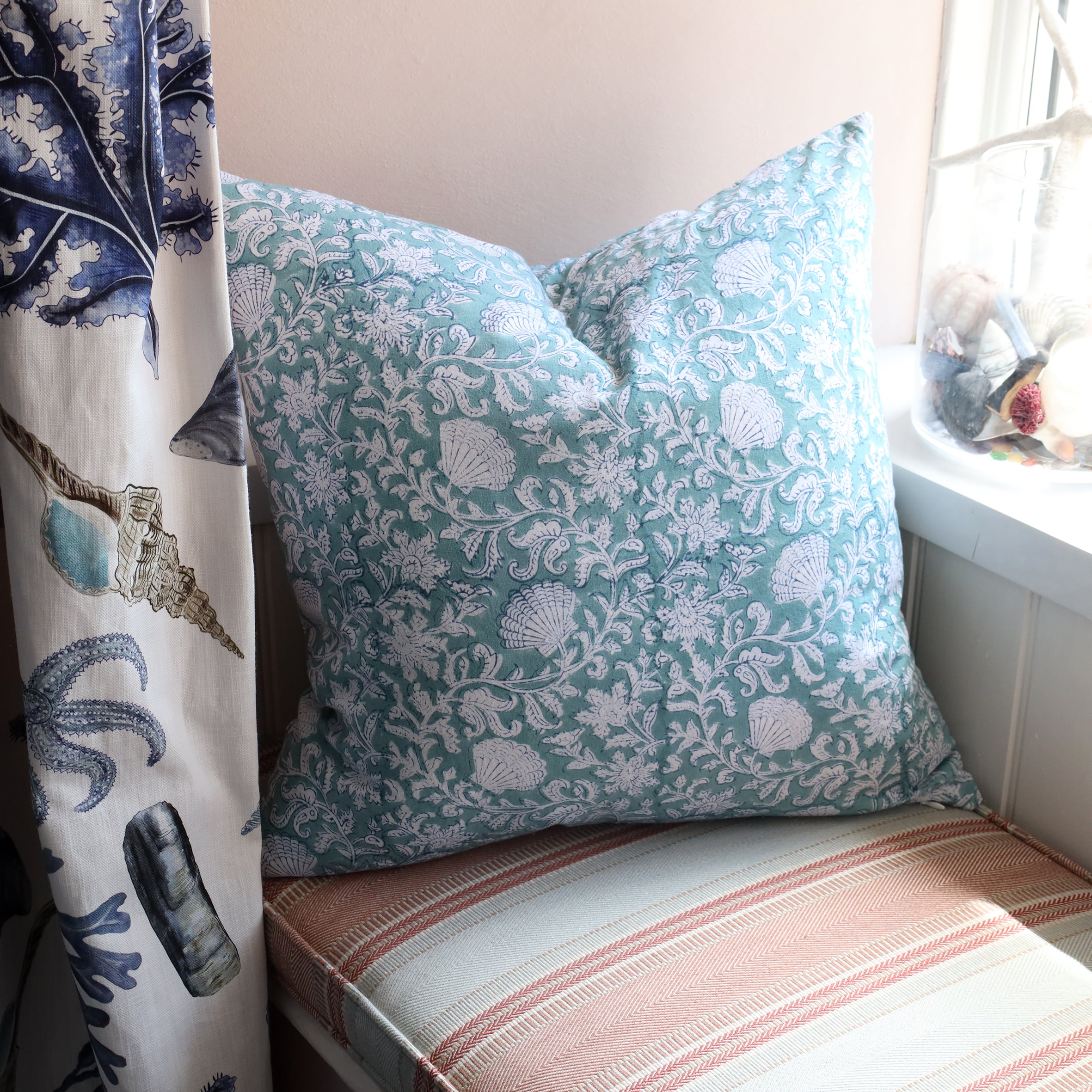 Sea Breeze Seashell Flower cushion which is Hand block printed fabric in a turquoise blue .Cushion is on a window seat setting with a curtain made in our Rockpool fabric and a jar of shells on the window ledge.