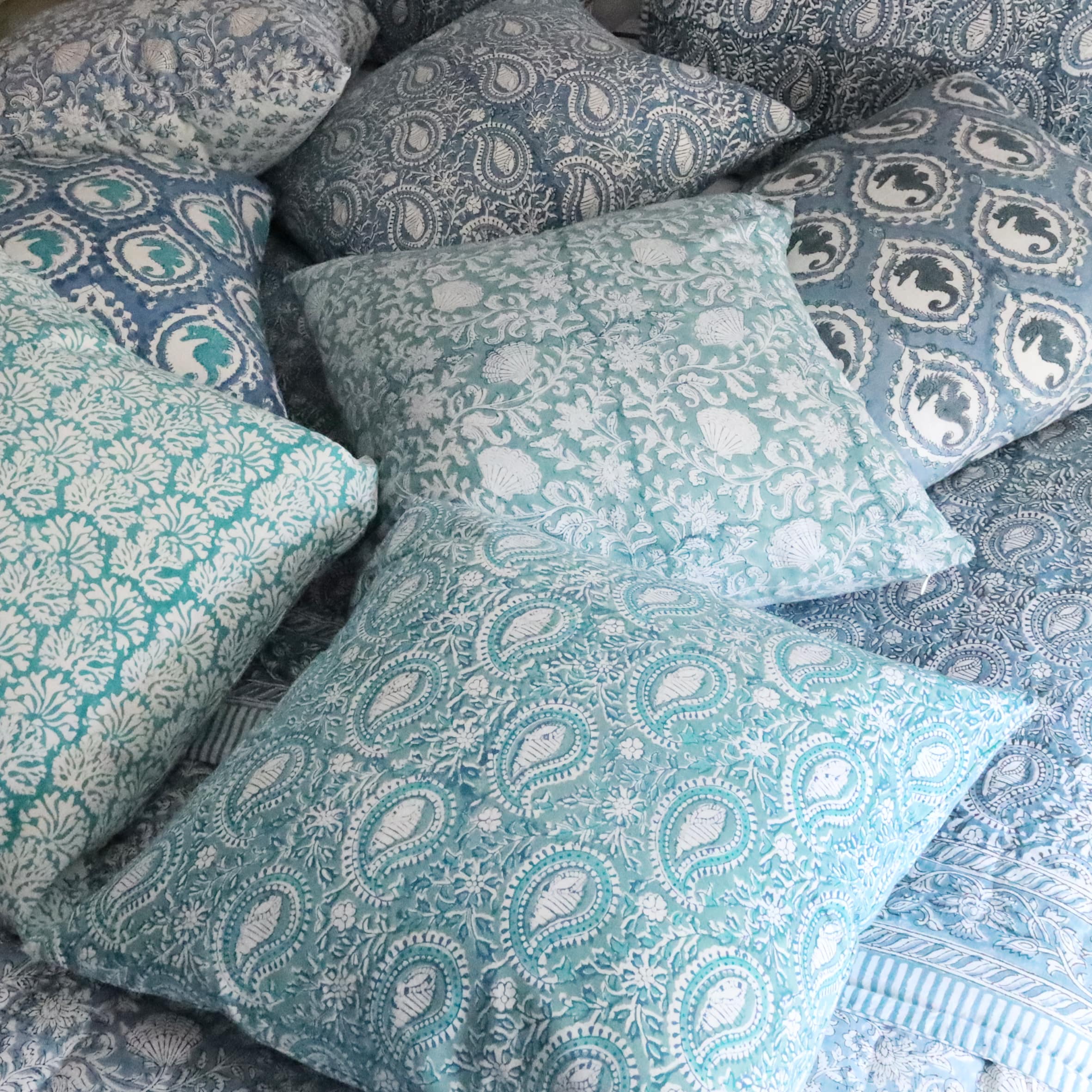 Sea Breeze Seashell Flower cushion which is Hand block printed fabric in a soft blue and the print is swirling shells and tendrils in white on a pile of other Hand block printed cushions