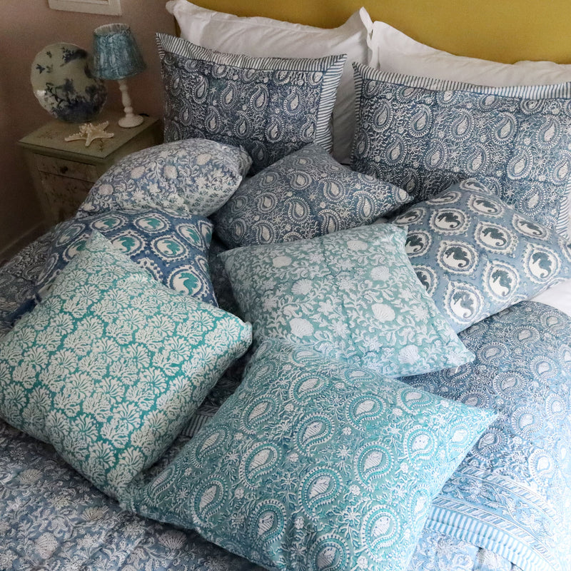 Coastal Blue Coraline cushion which is Hand block printed fabric in a soft blue and the print is soft turquoise blue with white coral print on a bed.Cushion is piled on a bed with lots of other hand blocked painted cushions Behind the cushions and pillows ia a mustard yellow Headboard with a bedside cabinet with various items on.