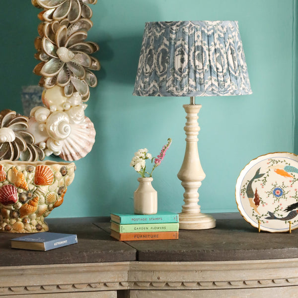 Medium Seahorse Cameo in Ocean pleated lampshade in hand blocked print on a wooden lampbase,placed on a sideboard.Also a small stack of coloured books,A plate on a stand and in the foreground a shell vase.Behind on the wall is a shell mirror 