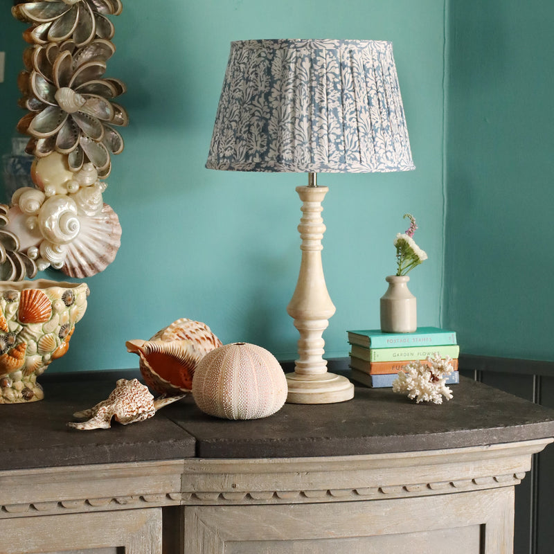 Medium Coastal Blue pleated lampshade on a wooden lampbase placed on a sideboard.On the sideboard are several shells and some small books.To the side you can see a shell vase and the edge of a shell mirror
