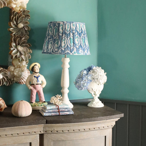 Large Seahorse Cameo pleated lampshade on a white lampbase on a sideboard.Also on the sideboard is a small white shell ornament,a pile of small notebooks and a sailor ornament,on the wall behind is a shell mirror