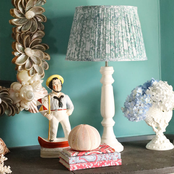 Large Seabreeze Seashell Flower pleated lampshade in hand blocked print on a white lampbase.Also on the sideboard is a small white shell ornament,a pile of small notebooks and a sailor ornament,on the wall behind is a shell mirror
