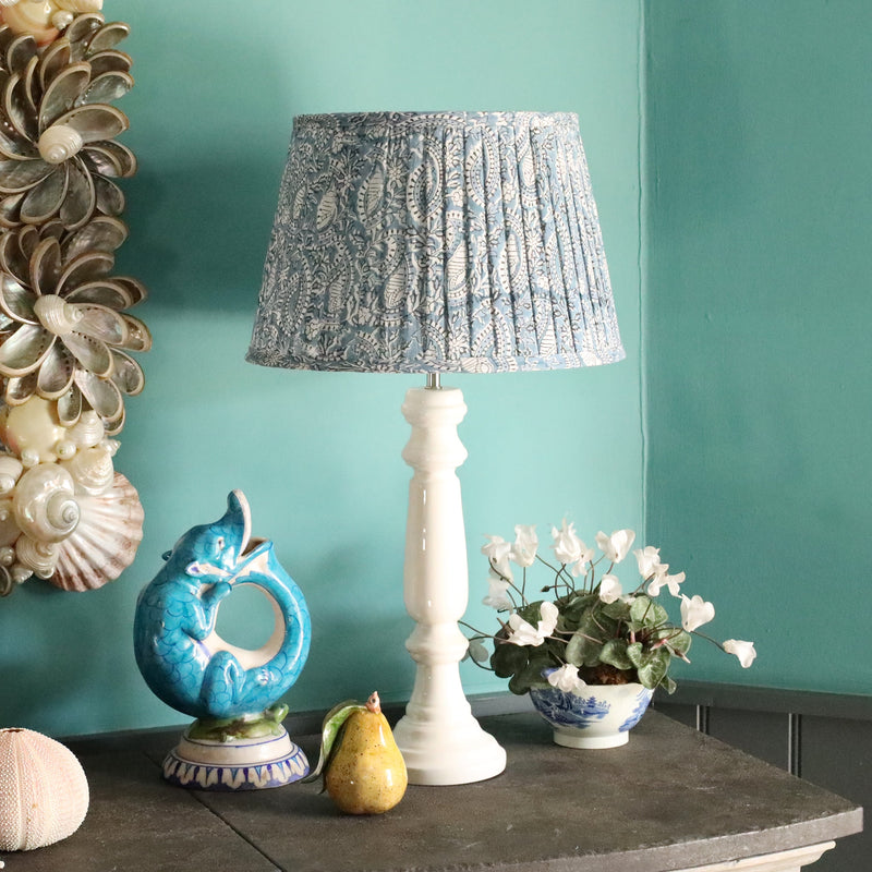 Large Azure paisley pleated lampshade on a white lampbase on a sideboard.Also on the sideboard is a small white plant in a blue and white pot,a ceramic pear and a jug shaped like a fish,on the wall behind is a shell mirror