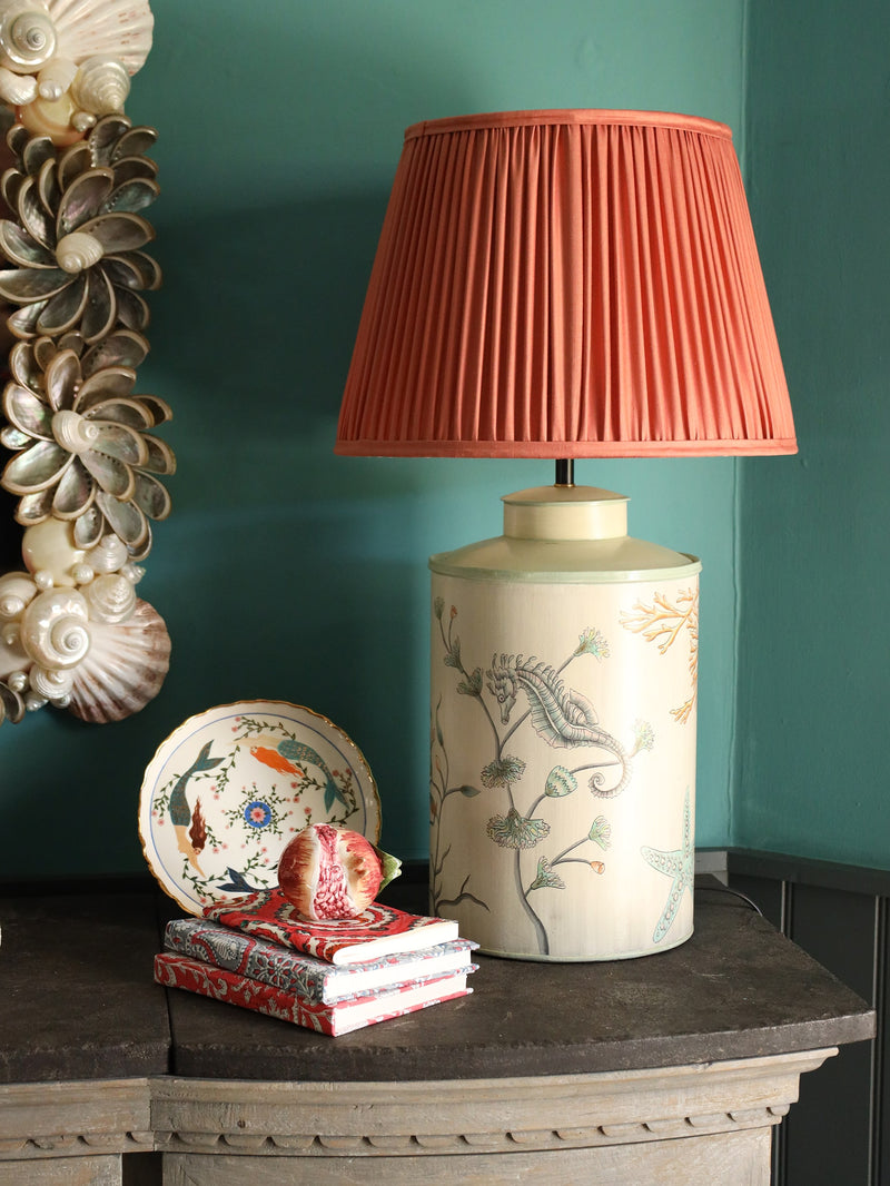 Hand Block printed paper backed notebooks stacked on a shelf with ceramic fruits in the background. On the shelf is our hand painted lampbase with a pleated lampshade,to the left is a shell mirror on the wall.