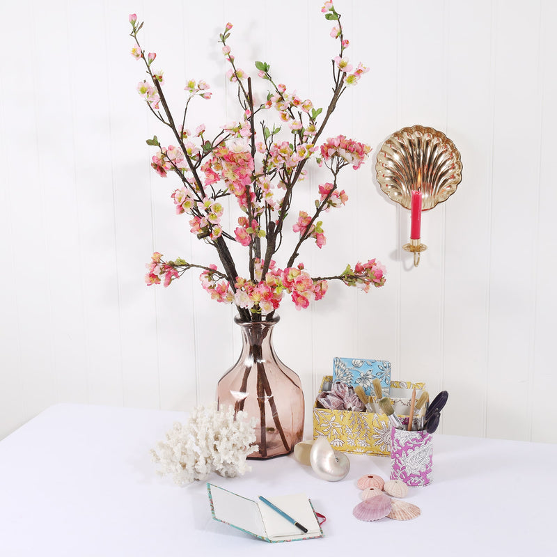 summer still life scene of pink glass vase filled with pink summer blossom branches, soft pastel coloured stationery and shells with a brass wall sconce and lit pink candle hanging on the wall above.