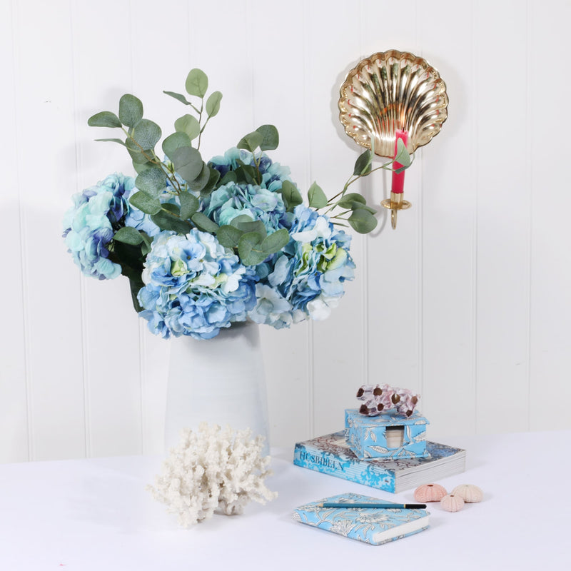 summer still life scene of white jug filled with hydrangeas and eucalyptus, soft blue stationery and shells with a brass wall sconce and lit pink candle hanging on the wall above
