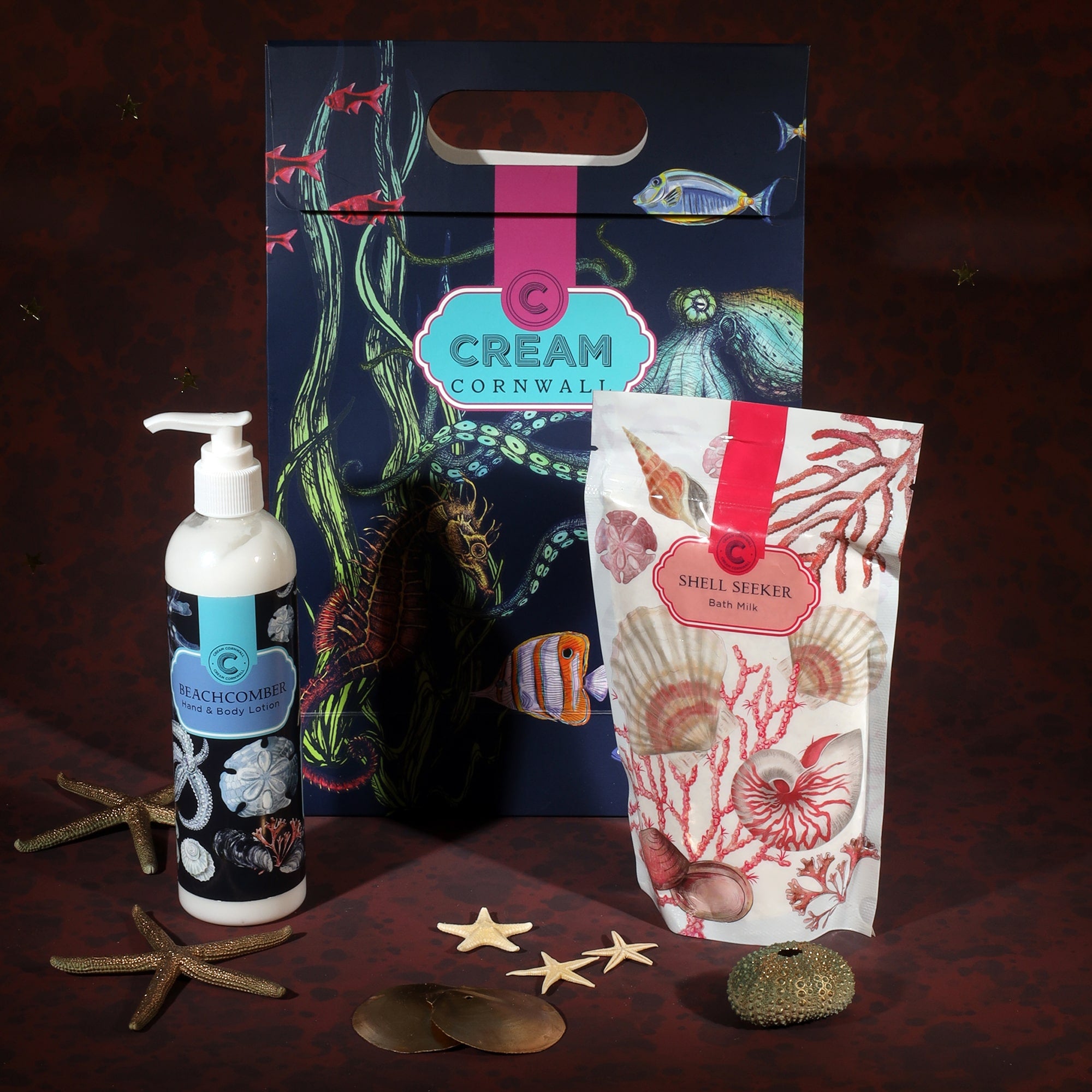 A Cream Cornwall gift set featuring Beachcomber hand and body lotion and Shell Seeker bath milk. The products are displayed with a colorful gift bag decorated with marine life, including fish, seaweed, and a seahorse. Decorative starfish and seashells are scattered around the items, enhancing the coastal theme against a deep red background.