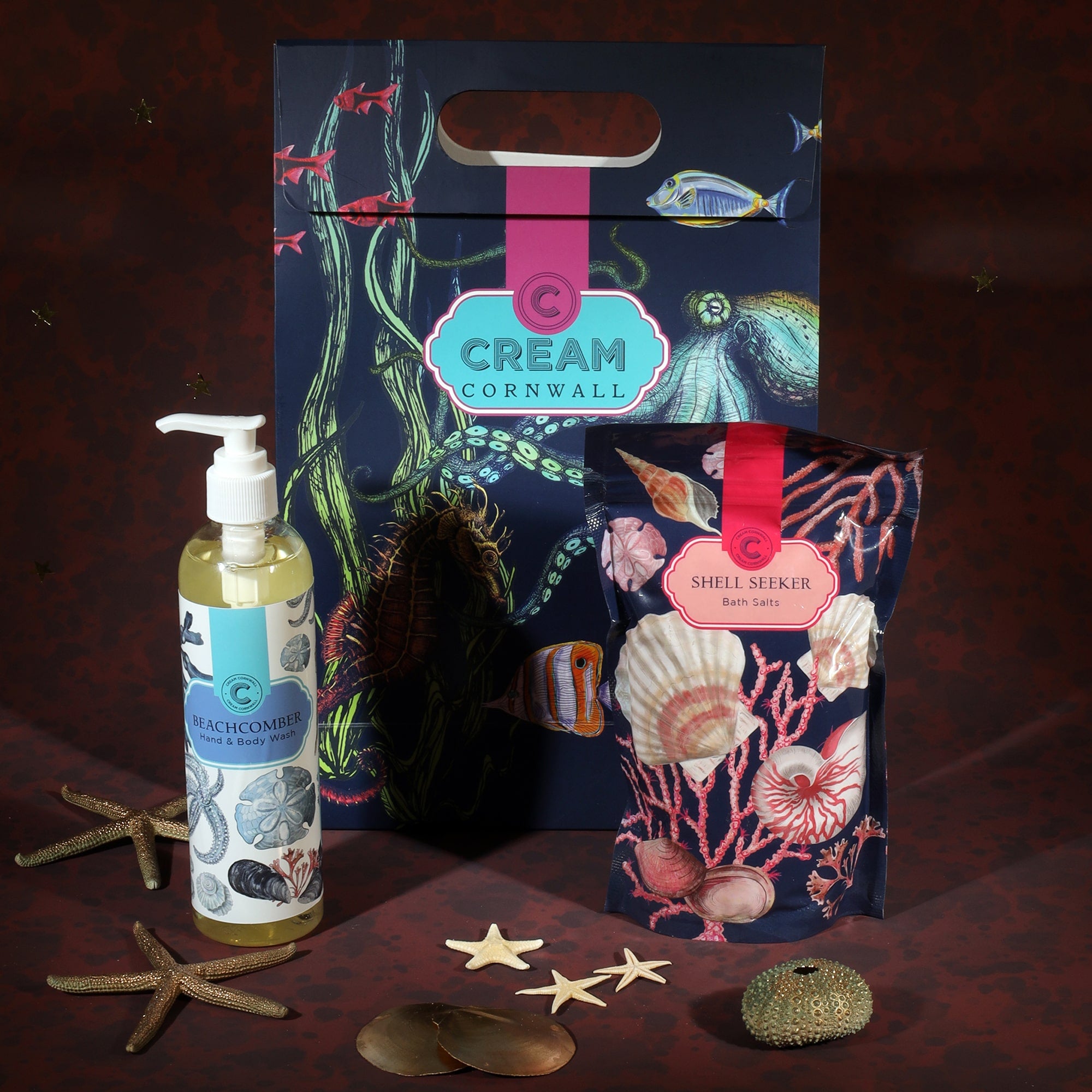 A Cream Cornwall gift set featuring Beachcomber hand and body wash and Shell Seeker bath salts. The products are displayed with a vibrant marine-themed gift bag decorated with fish, seaweed, and a seahorse. Surrounding the items are decorative starfish and seashells, complementing the coastal theme against a deep red background