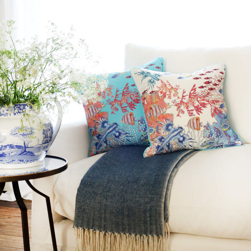2 cushions an  a white sofa with brightly coloured tropical underwater scene illustration on with the sun streaming through the window at the back and a large willow pattern jug filled with cow parsley
