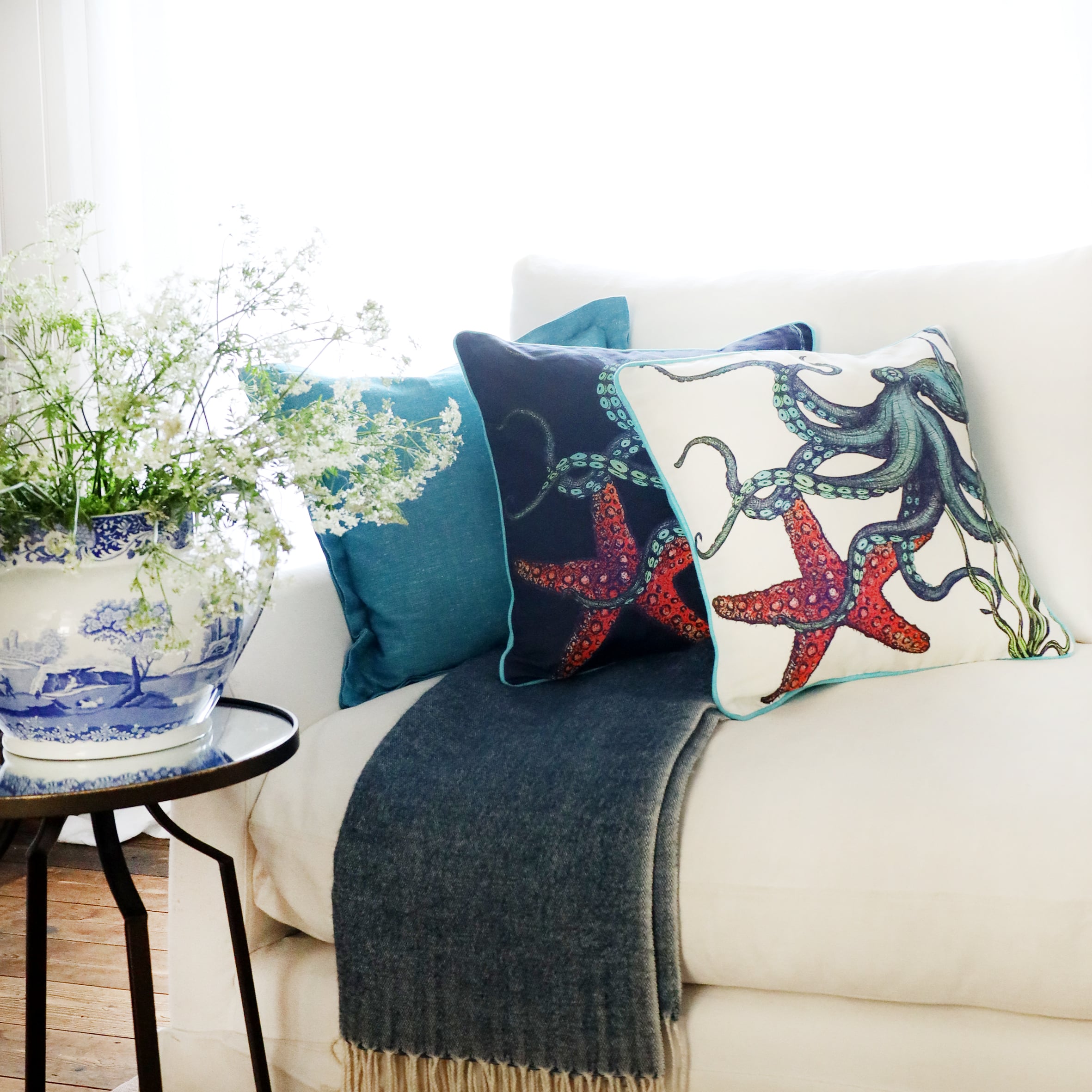 3 cushions an  a white sofa with brightly coloured octopus and starfish illustration on with the sun streaming through the window at the back and a large willow pattern jug filled with cow parsley