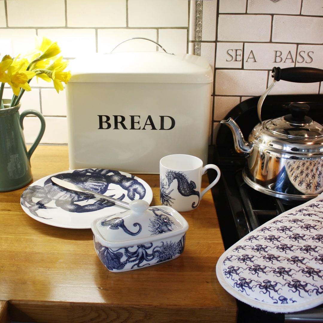 Butter dish in our Classic range,with seahorses,shells,octopus and other sea themed designs all over the base and the lid,placed on a wooden table in front of a bread bin with other matching classic tableware.Next to the table is an aga with a chrome kettle and  cream cornwall oven gloves