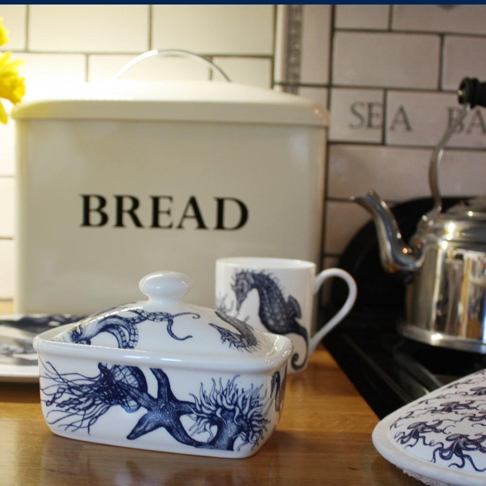 Butter dish in our Classic range,with seahorses,shells,octopus and other sea themed designs all over the base and the lid,placed on a wooden table in front of a bread bin with other matching classic tableware.Next to the table is an aga with a chrome kettle and cream cornwall oven gloves