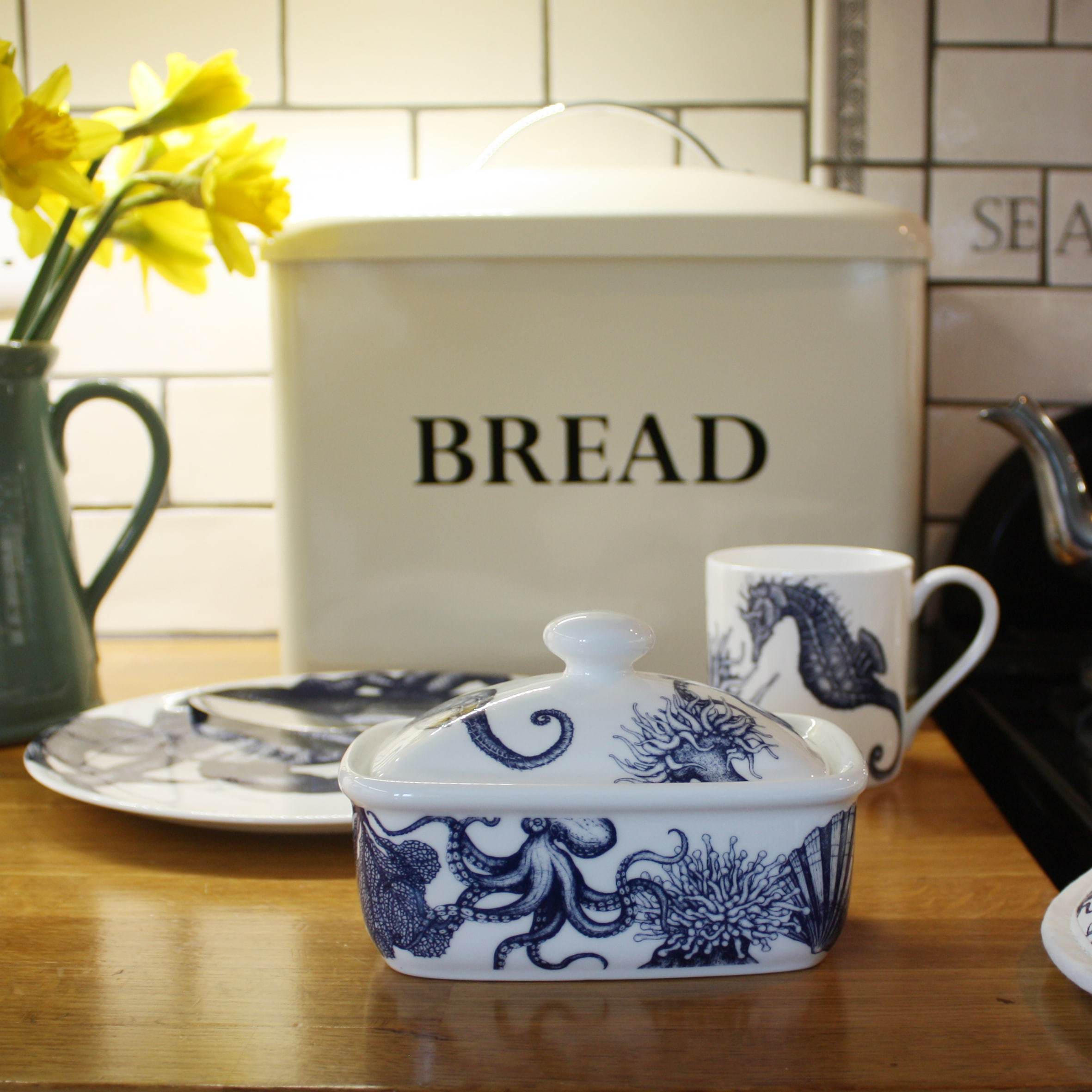 Butter dish in our Classic range,with seahorses,shells,octopus and other sea themed designs all over the base and the lid,placed on a wooden table in front of a bread bin with other matching classic tableware.
